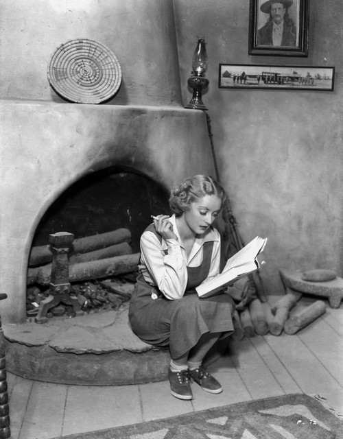 Bette Davis Seated Near the Fireplace with Cigarette on Hand while Reading a Book in White Long Sleeve and Apron Dress Photo Print - Item # VARCEL692867
