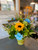 "Gone Fishin'" captures the vibrancy of a summer day. A bold sunflower acts as the radiant sun, surrounded by a sea of bright blue carnations. Nestled among the blooms, a whimsical fake fish peeks out, lending a playful charm. This lively arrangement, exuding warmth and amusement, is a perfect nod to leisurely fishing trips under the sunny sky.
