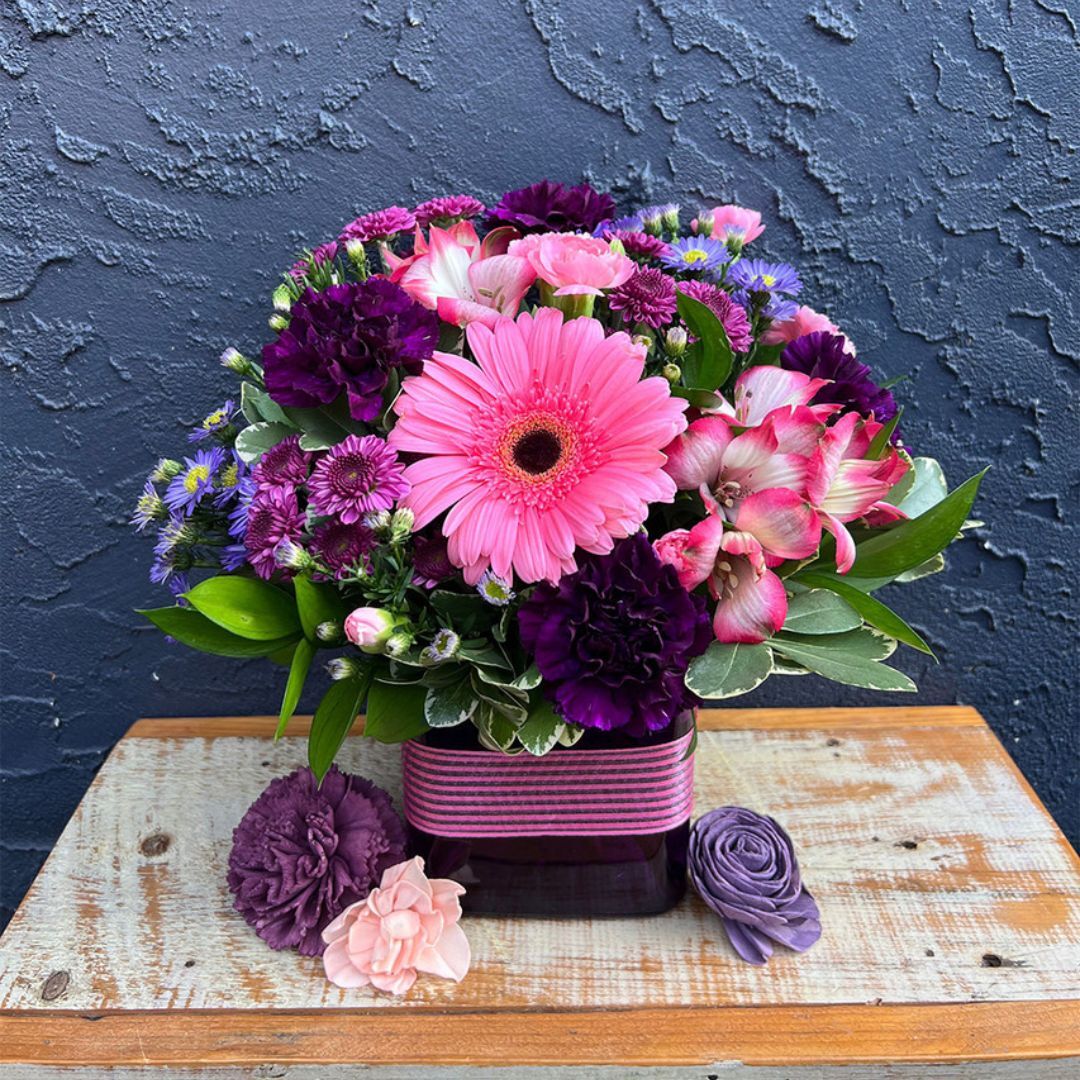 Pink gerbera with alstroemeria and purple mums in a ribbed purple vase on a wood table, dark textured wall behind