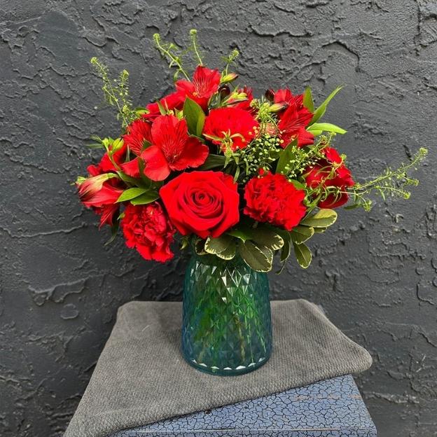 Compact red flower arrangement in a blue-green diamond-cut vase, with roses, carnations, and alstroemeria.