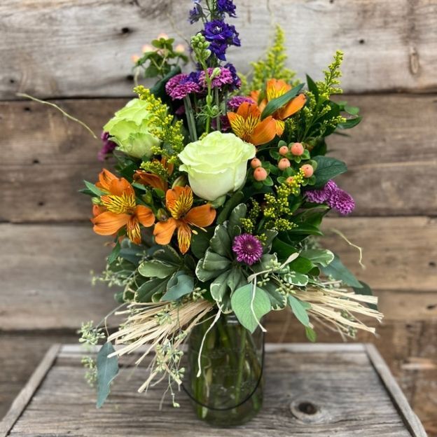 Rustic bouquet in a twine-tied mason jar with orange alstroemeria, purple blooms, and a pale green rose against wood.