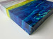 Photo of corner of canvas print of original abstract landscape painting, on canvas, with blues, green, yellow, gray, purple. White background around canvas.