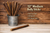 A rustic product photo of 12" medium bully sticks arranged in a weathered metal bucket on a light wood-textured surface. One bully stick lies in the foreground. Cream-colored overlay text reads: ‘12"Medium Bully Sticks’ with bullet points: ‘Very Low Odor,’ ‘All Natural,’ and ‘45-54g per stick.’ The background features darker wooden planks with visible grain and knots, while the lighter wood floor adds warmth and contrast, reinforcing the natural, handcrafted Mushyface brand aesthetic.