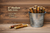 A rustic product photo of 6" medium bully sticks arranged in a weathered metal bucket on a light wood-textured surface. Two bully sticks lie in the foreground. Cream-colored overlay text reads: ‘6" Medium Bully Sticks’ with bullet points: ‘Very Low Odor,’ ‘All Natural,’ and ‘55–64g per stick.’ The background features darker wooden planks with visible grain and knots, while the lighter wood floor adds warmth and contrast, reinforcing the natural, handcrafted Mushyface brand aesthetic.