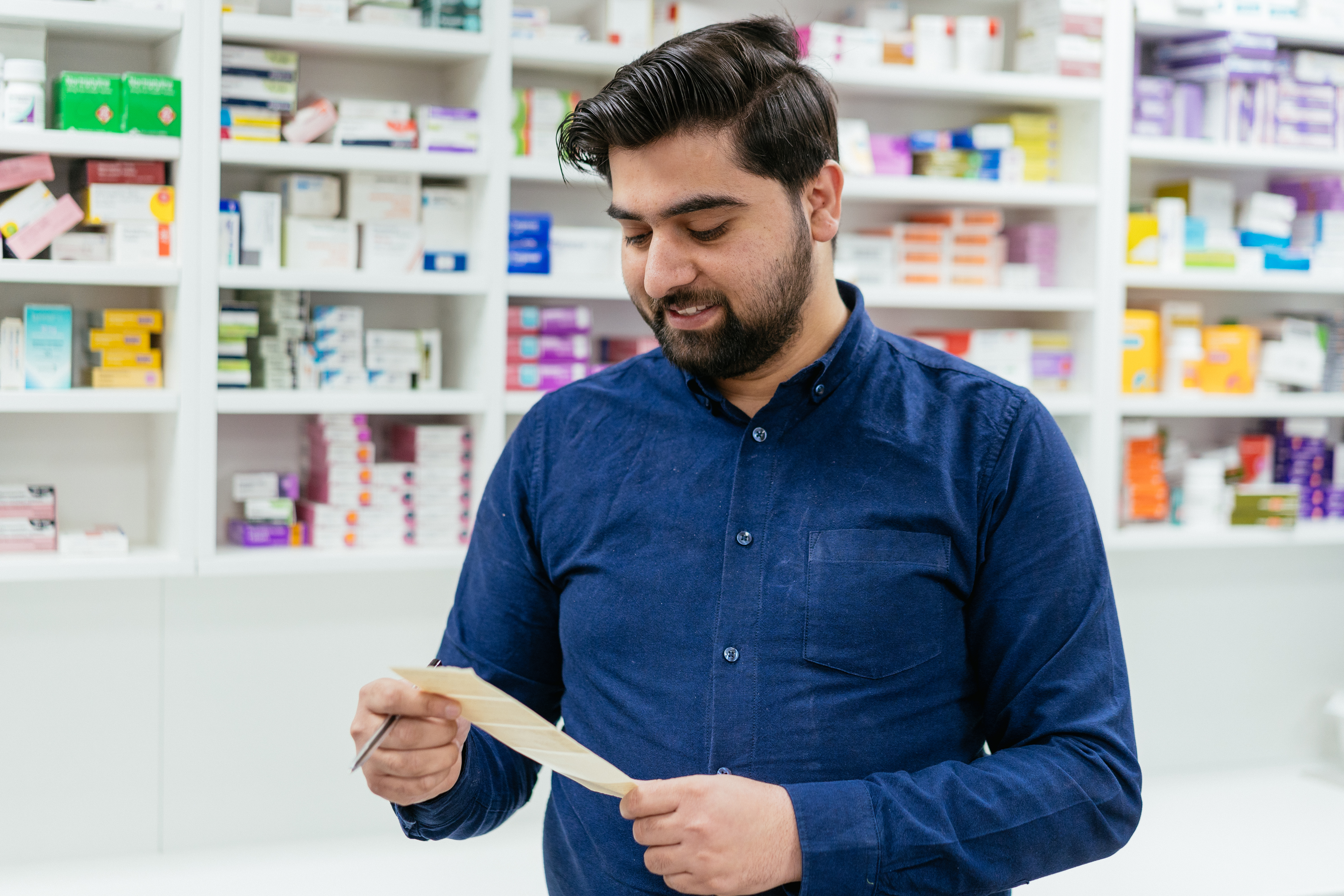 A Well Pharmacist checking a prescription in front of shelves  stacked with medicines.
