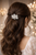 Woman seen from behind with long wavy dark hair and a small silver flower-shaped accessory, warm lights behind