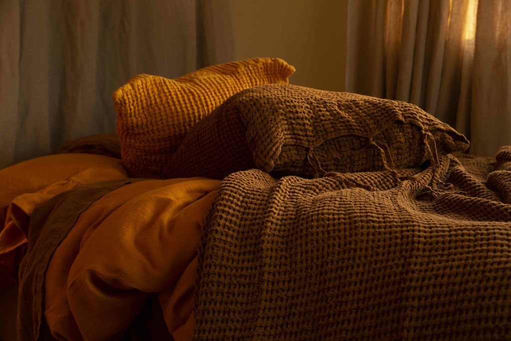 Close up view of two linen pillows laying next to one another on a bed of ruffled linen sheets- one dressed in a saffron yellow pillowcase the other in waffle textured, cinnamon pillowcase.