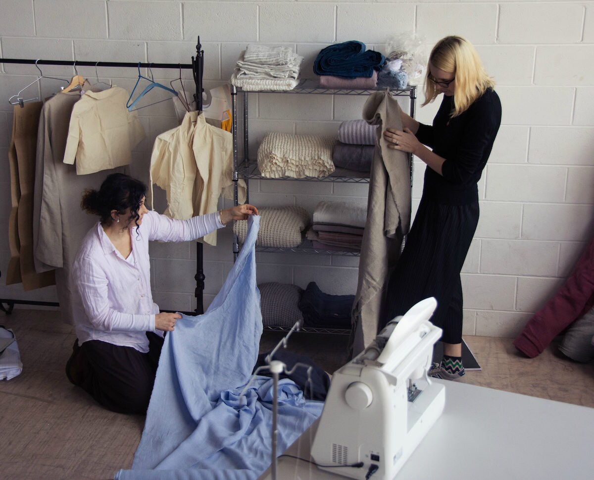 Image of House of Baltic Linen owner with daughter sorting through linen fabric in their studio. Studio features a rack full of calico toiles, a sewing machine, an overlocker on the table and an industrial shelving unit for linen fabric scraps and products.