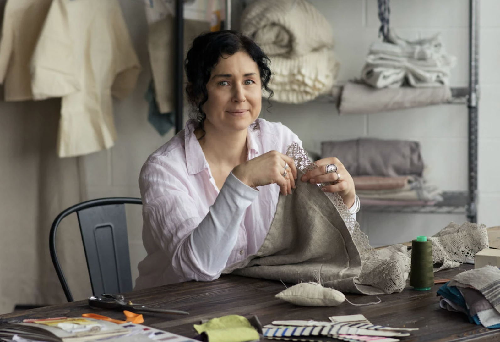 Photograph of House of Baltic Linen owner sitting at her work table smiling at the camera whilst working on linen lace trim of a pillowcase in her hands.