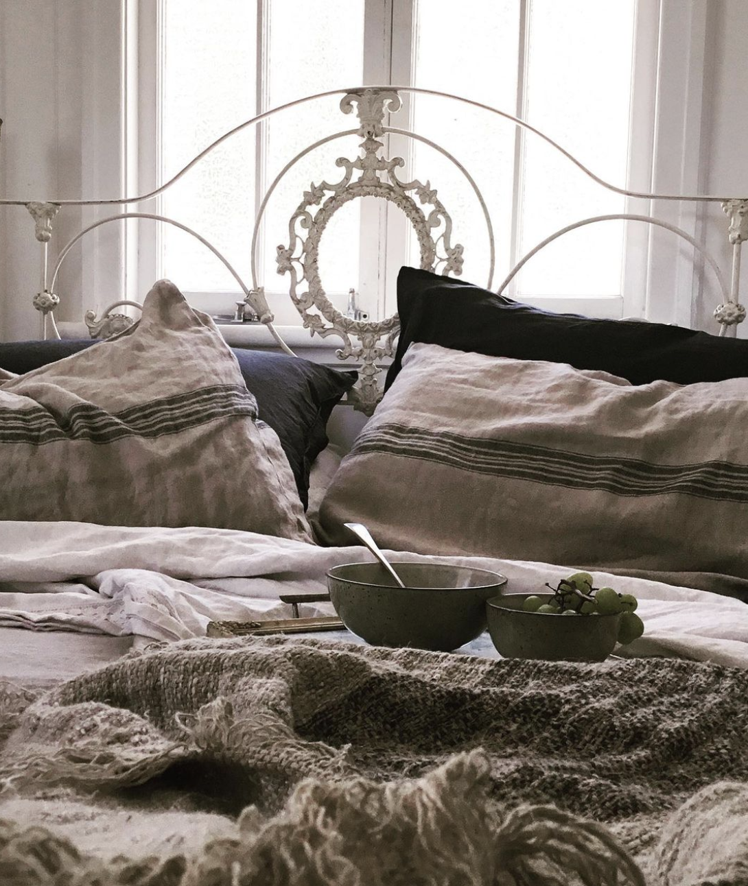 Close up of two striped linen pillows next to each other with a ceramic bowl, spoon and grapes on a tray in the foreground placed on the bed.