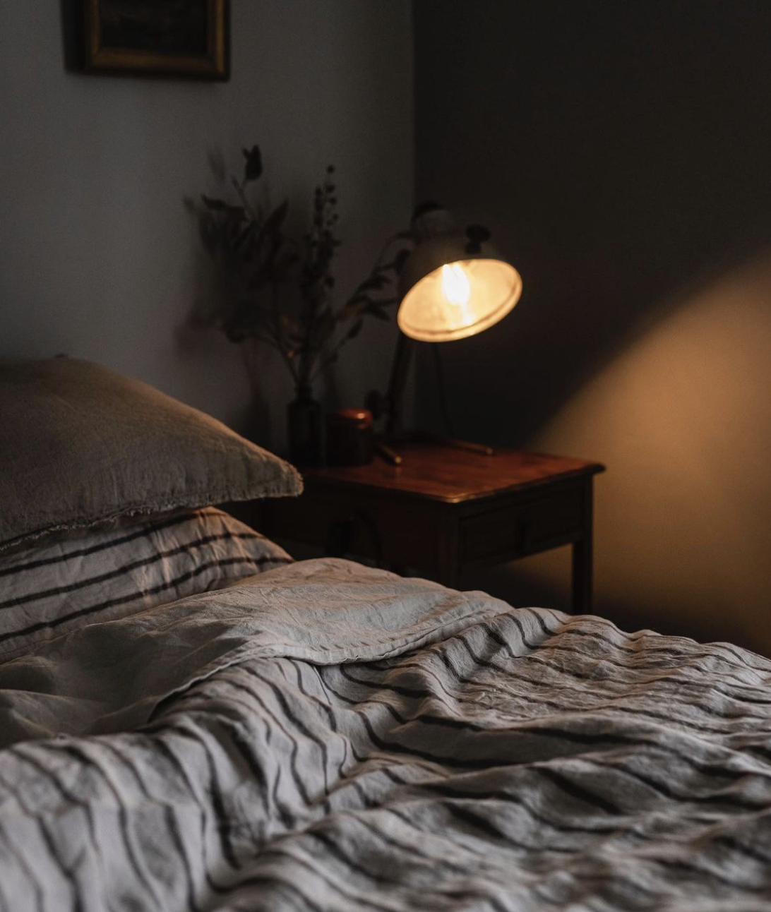 Moody photograph of a close up of a bed in striped black and grey linen sheets with a bedside table and a lit up lamp next to it.