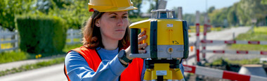 Construction worker in hard hat and high-visibility vest setting up a Leica Rugby 60DG dual grade laser on a tripod at a roadworks site