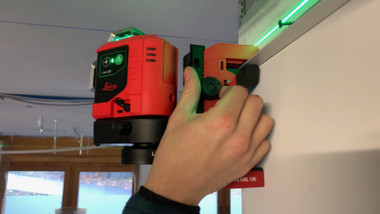 Hand fitting a Leica Lino L6G on a UAL 130 wall-mount adapter along a drywall track with a green line on the ceiling edge