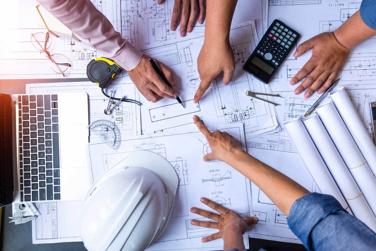 Three engineers collaborating over technical drawings and specification documents, with hands pointing to plans surrounded by a laptop, hard hat, calculator, tape measure, drawing compass, pens, and glasses on a work surface
