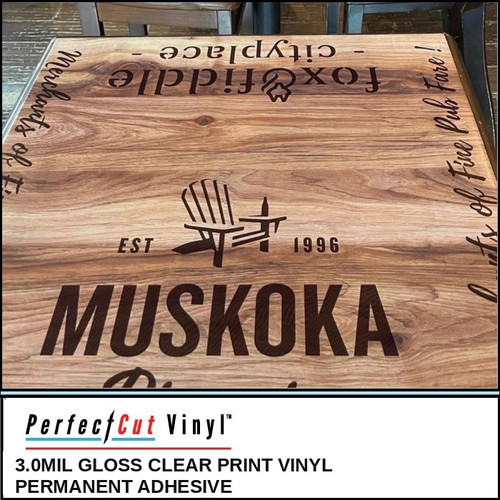 PerfectCut Vinyl 3.0mil gloss clear print vinyl with permanent adhesive, shown as a branded "Muskoka Est. 1996" logo graphic applied directly to a wood-grain restaurant table surface.