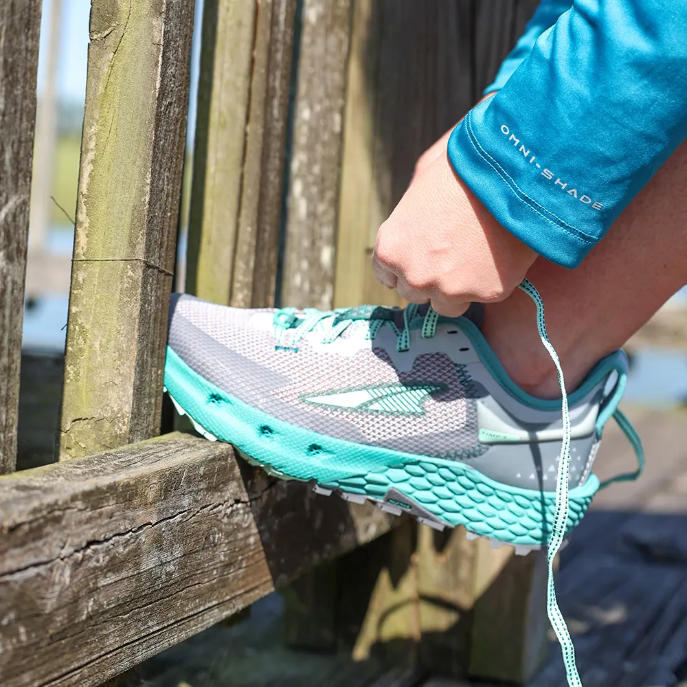 Womens with foot on a wooden fence tieing her laces of her teal and grey Altra Women's Torin 5 Luxe Sneaker.