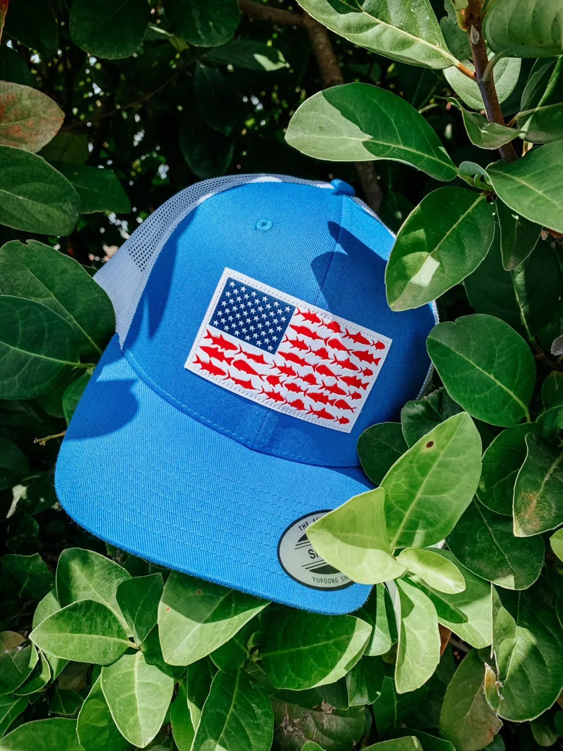 A blue Columbia PFG Fish Flag Hat resting in a green bush.