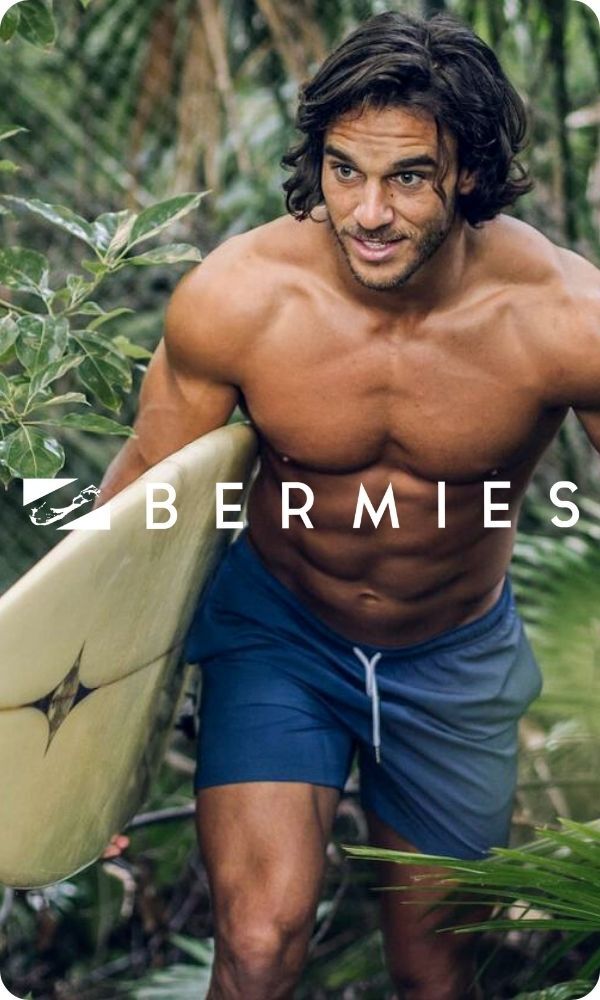 Man walking with a surfboard wearing Bermies swimwear, logo on top