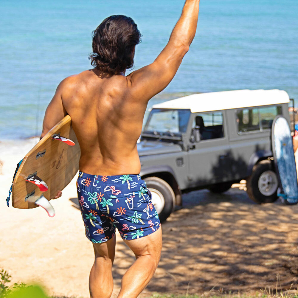 Man holding a surf board and waiving while wearing Chubbies Neon Lights Stretch Boardshorts