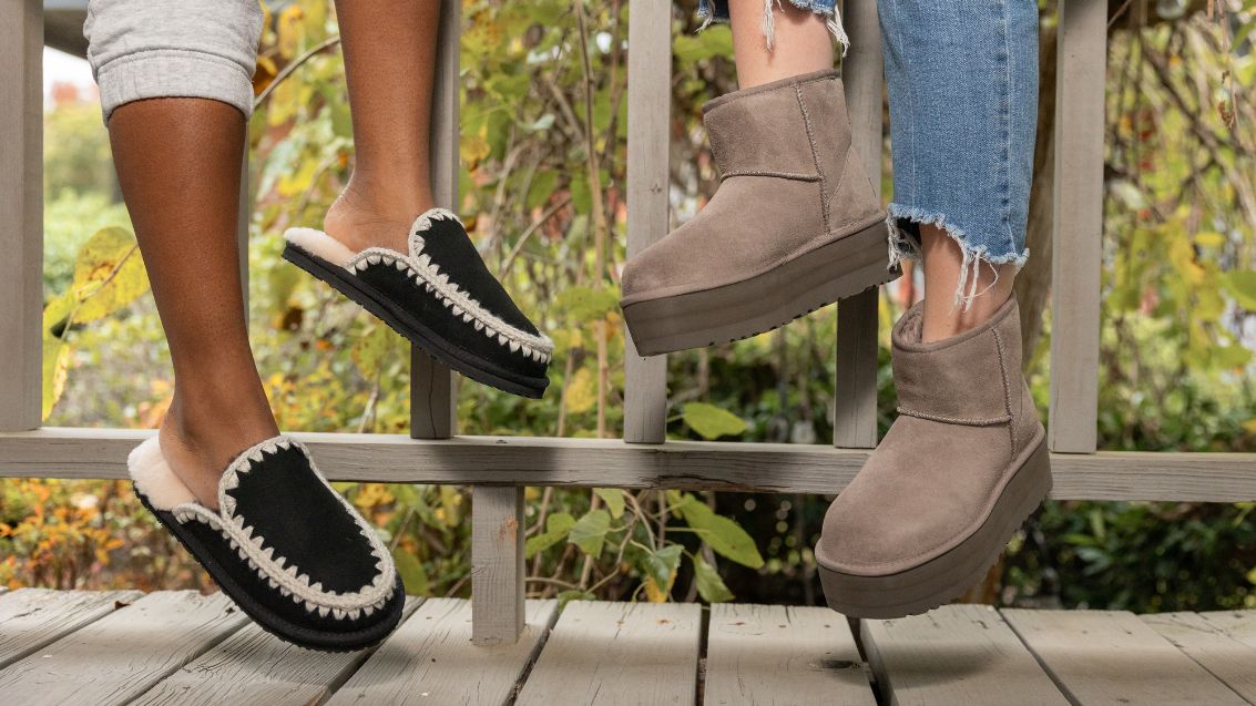 Two women with their feet dangling off of a railing. One wearing comfy Mou slippers and one wearing trendy Ugg boots,