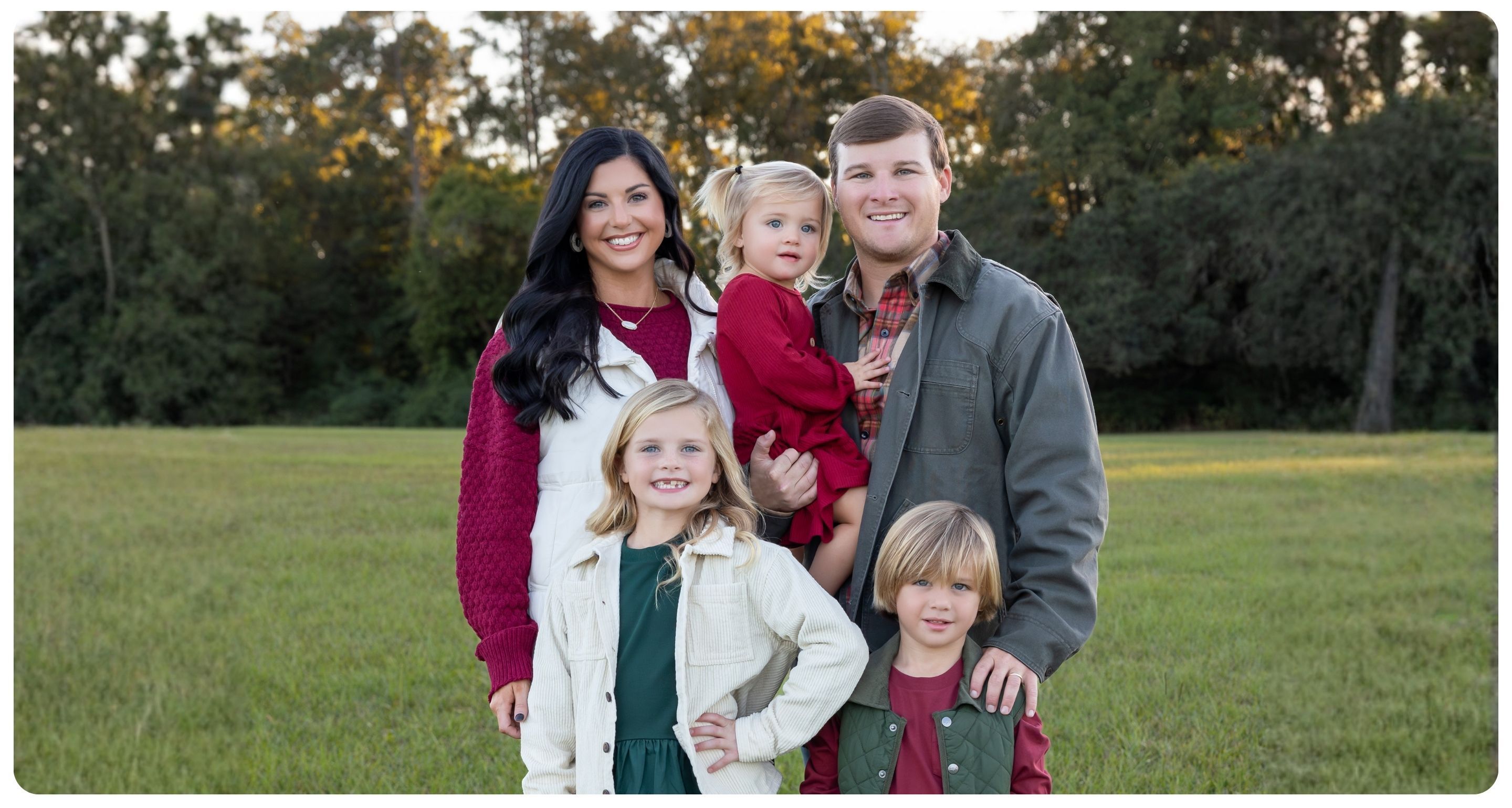 Family with three kids standing in a field with trees behind them dressed in Christmas color apparel.