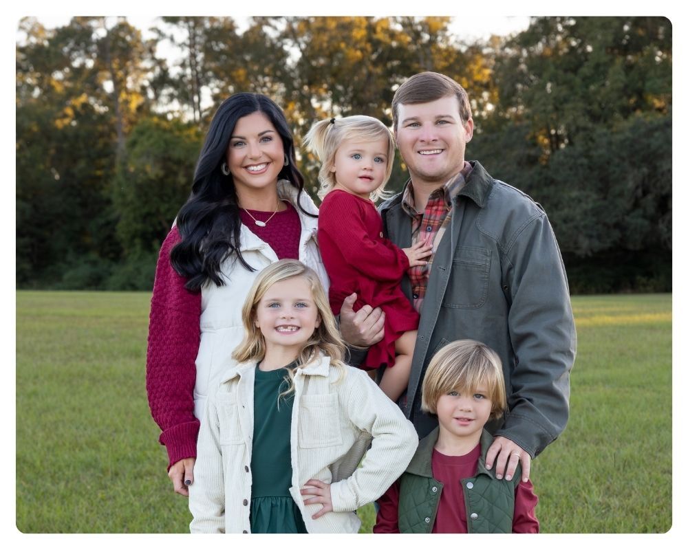Family with three kids standing in a field with trees behind them dressed in Christmas color apparel.