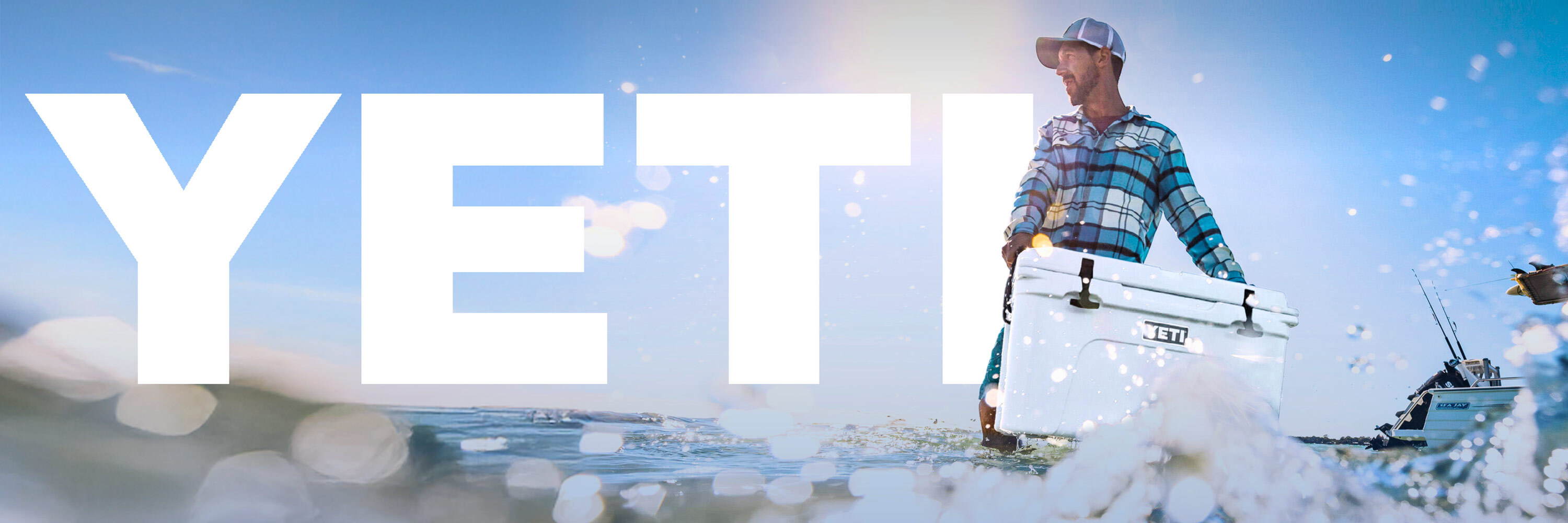 Man carrying a YETI cooler in shallow water. YETI logo behind man over the blue sky.