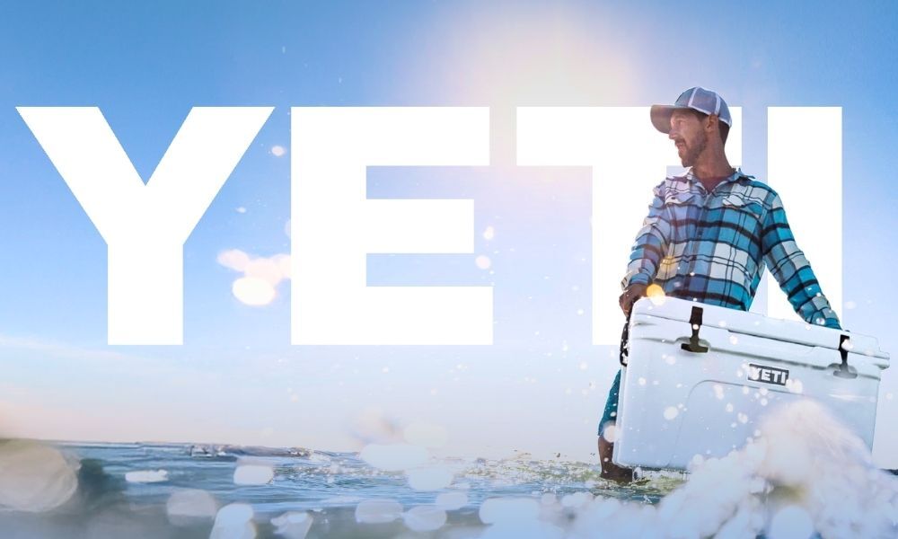 Man carrying a YETI cooler in shallow water. YETI logo behind man over the blue sky.