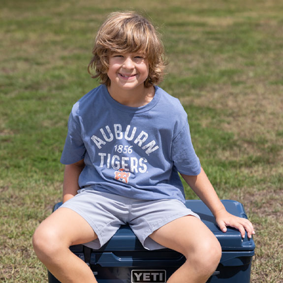 Shop Collegiate Kids. Boy sitting on a Yeti cooler outside wearing a blue Auburn Tigers t-shirt.