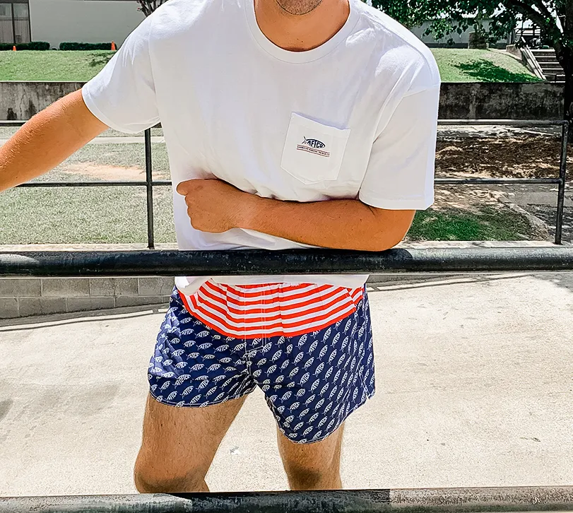 Close-up picture of a man wearing Aftco logo red, white and blue swim trunks and an Aftco white pocket t-shirt.