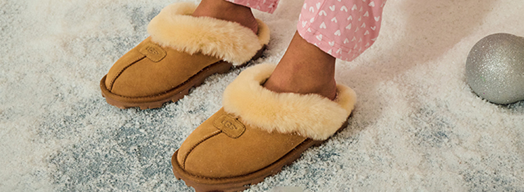 Close up of a woman's feet wearing a pair of tan Ugg slippers and pink and white heart pajamas.