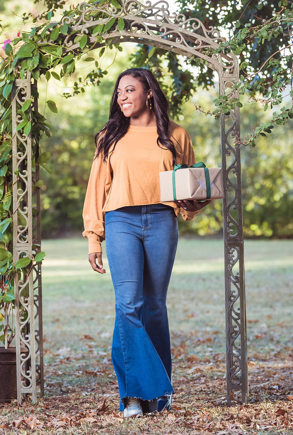 Woman outside in a garden holding a gift and wearing flare jeans and long sleeve light rust colored blouse.