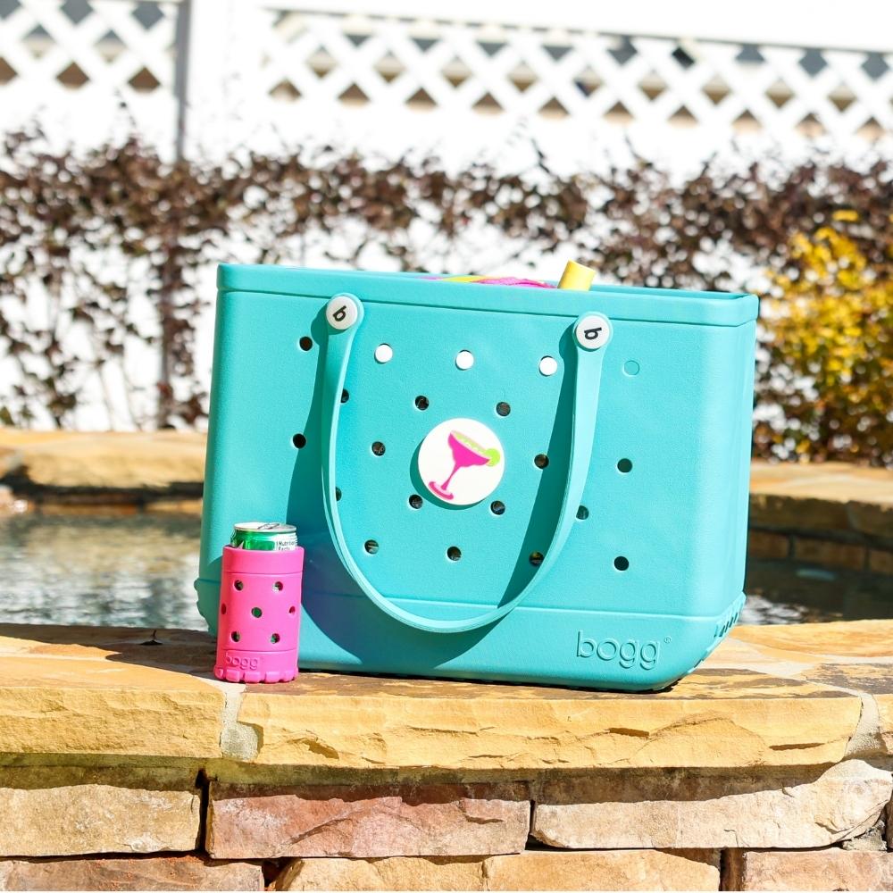 Turquoise Bogg Bag at the edge of a pool with a Margarita Bogg Bit on the bag and a drink inside a pink Bogg Boozie in front of the bag.