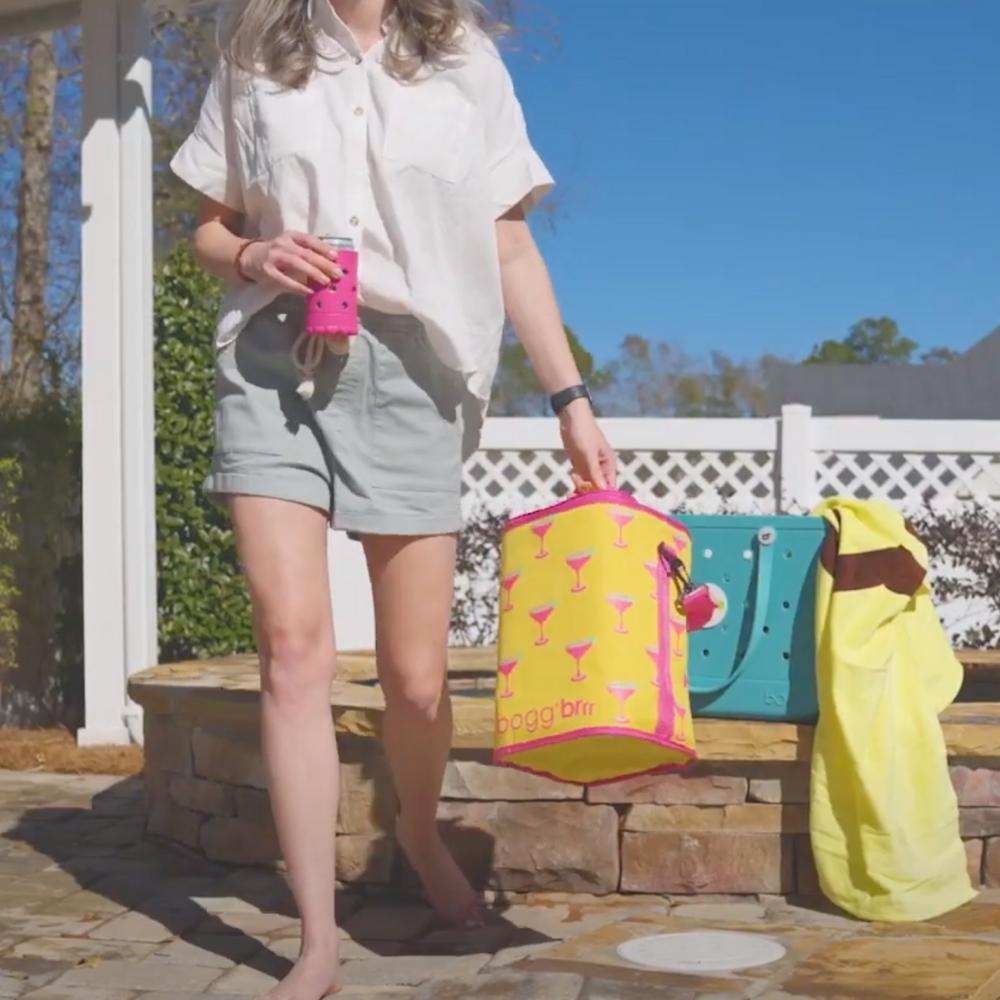 Woman holding a can with a pink Bogg Boozie and walking holding a Margarita Brrr Half Cooler insert. Large Turquoise Bogg Bag and beach towel sitting behind her.