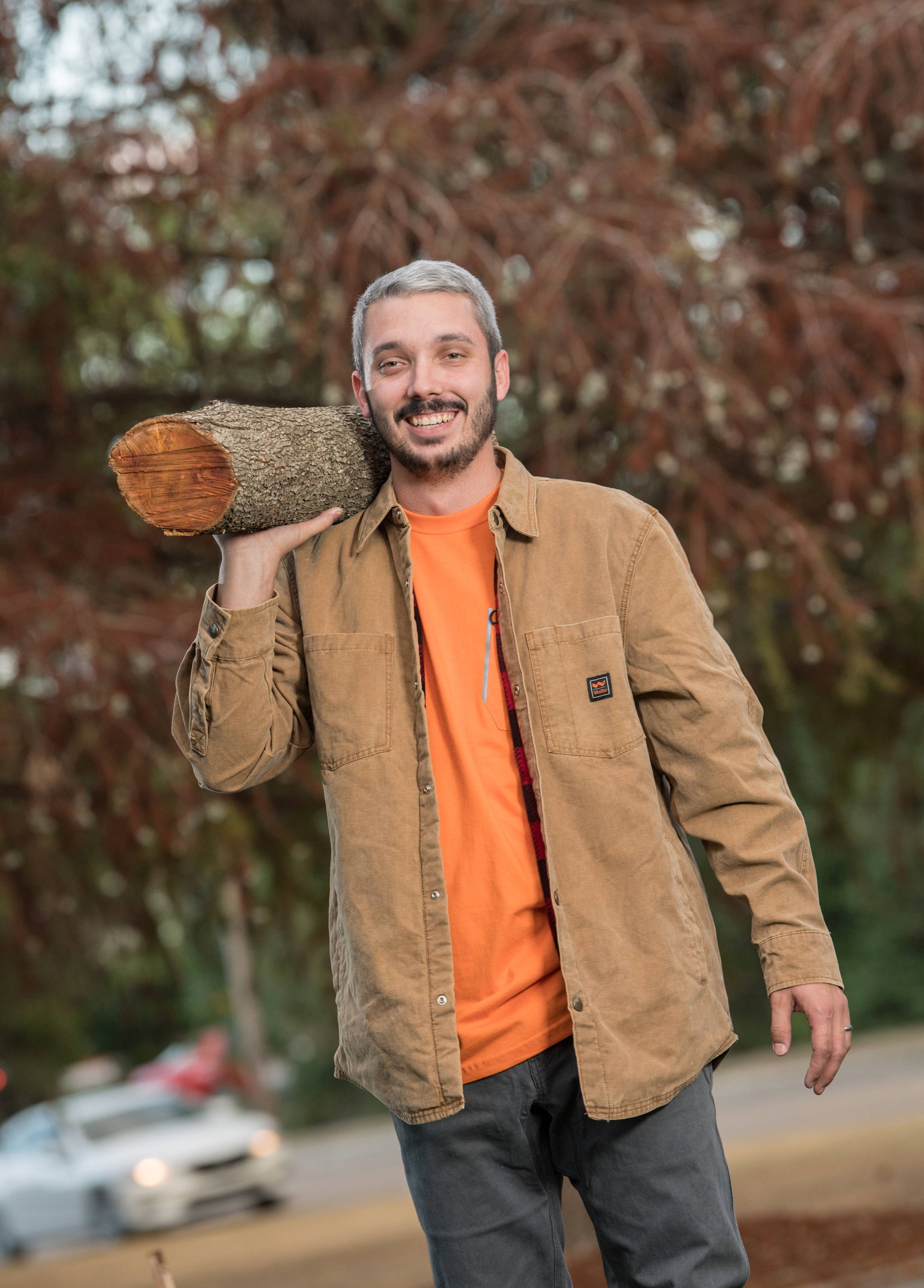 Man outside carrying a log wearing an orange Carhartt t-shirt and a tan long sleeve Carhartt button down work shirt.