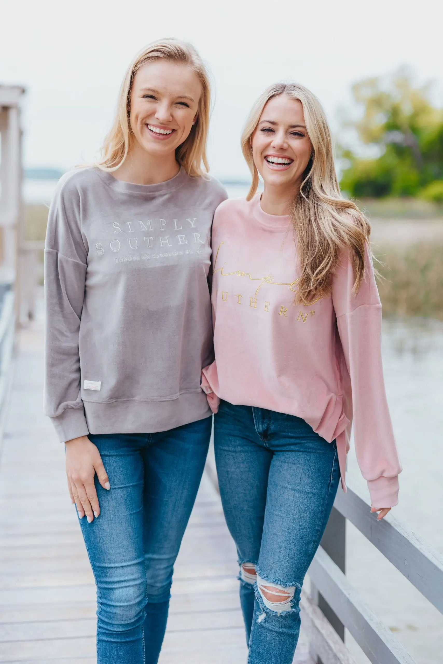 Two girls standing on a dock on a bay wearing a grey and a pink Simply Southern pullover.