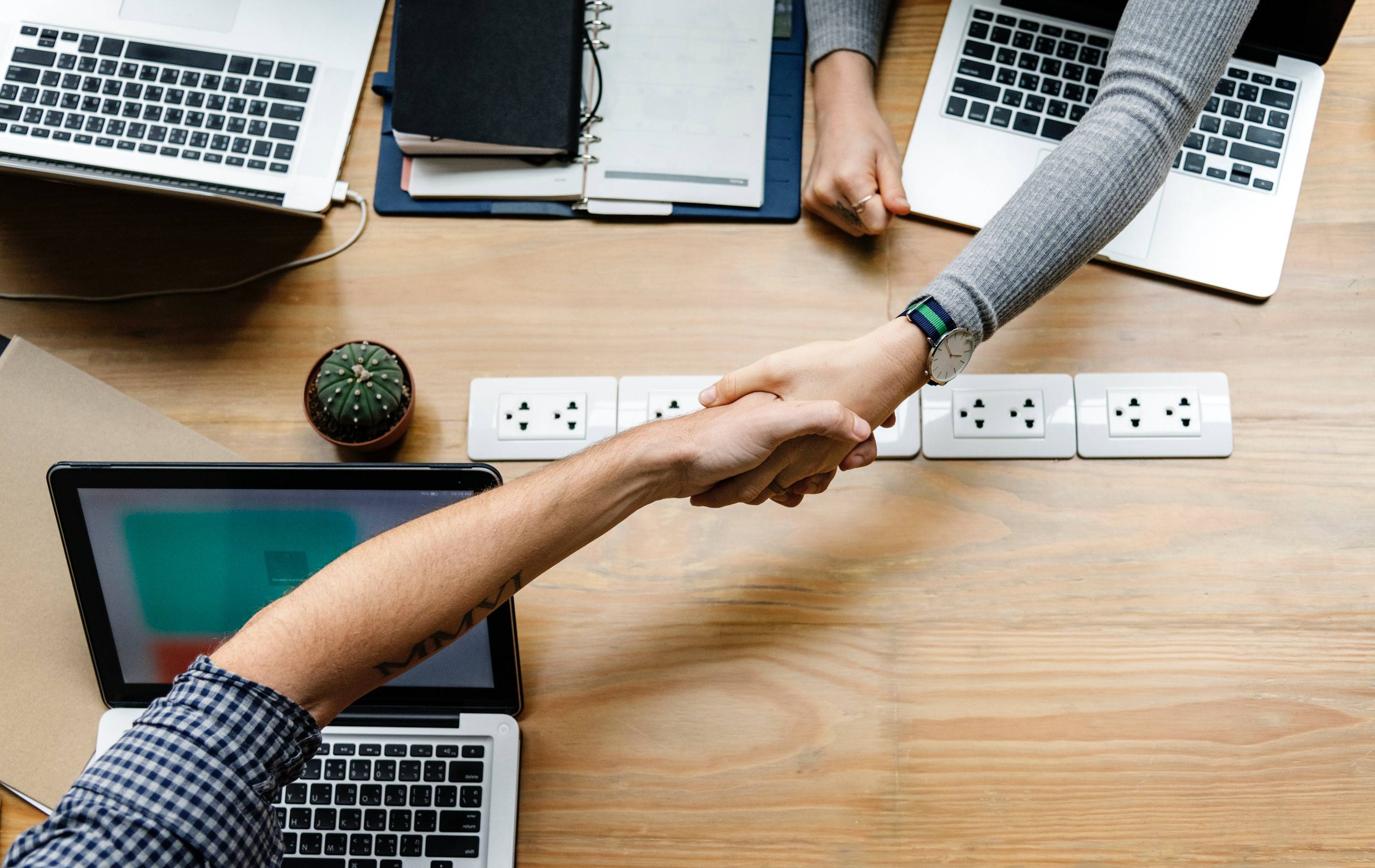 Man and woman shaking hands at a work table over their laptops.