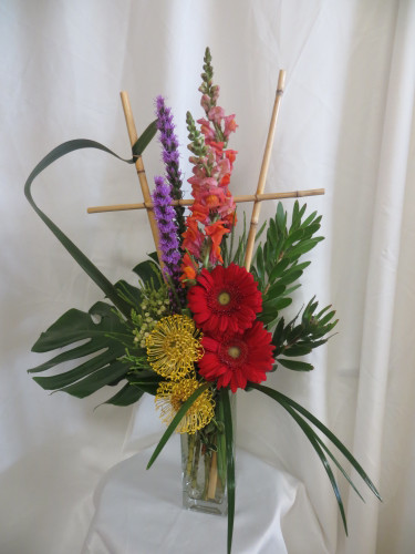 Perfectly Pincushion Protea Exotic Flower Arrangement by Enchanted Florist. This unique bouquet of tropical flowers includes red gerbera daisies, purple liatris, orange snapdragons and pincushion protea in a tall clear vase. Them the bouquet is accented with folded leaves and a bamboo trellis for a unique look. SKU RM169