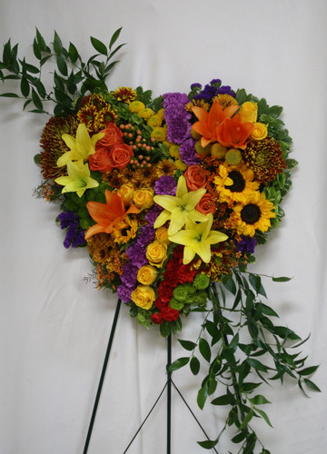Garden of Memories Yellow, Orange, and Purple Heart Wreath by Enchanted Florist Pasadena TX. A vibrant patchwork of beautiful flowers including orange and yellow asiatic lilies, yellow sunflowers, orange and yellow roses, bells of ireland, green and yellow buttons, bronze mums, orange hypericum all arranged on a solid heart shaped wreath. Approximately 26"H x 20"W (size does not include stand)  
SKU RM551
