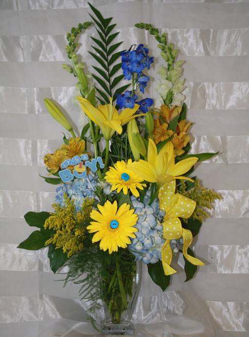 Cute As A Button New Baby Boy Flower Arrangement from Enchanted Florist. A perfect vase of mixed flowers to celebrate that special new bundle of joy. Includes blue hydrangeas and delphinium, yellow lilies & gerberas, and other flowers and is accented with adorable buttons to celebrate your new arrival. SKU RM324