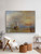 A large horizontal 60x44-inch matte fine art print of J.M.W. Turner's The Fighting Temeraire hanging in a home library. The archival landscape print features a dramatic, blazing sunset profile with rich gold and burnt orange tones casting a warm glow across a dark, moody wall.