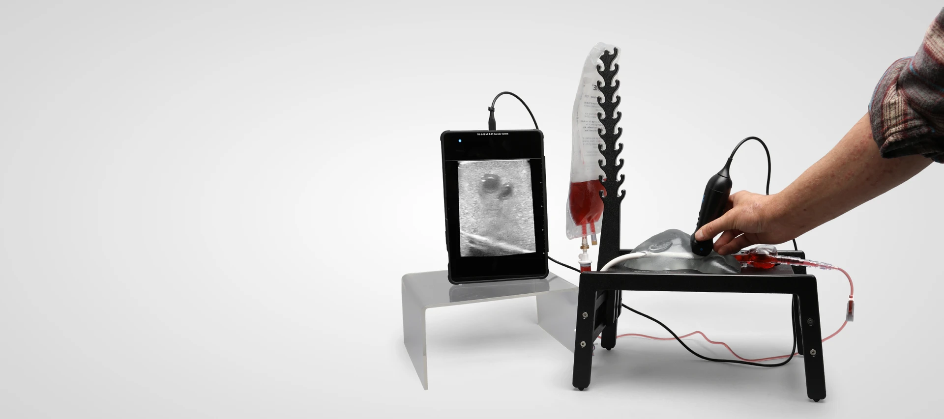 Clinician scanning a Sawbones neonatal IJ and carotid cannulation trainer with an ultrasound probe, with a blood-filled fluid bag and a tablet display.